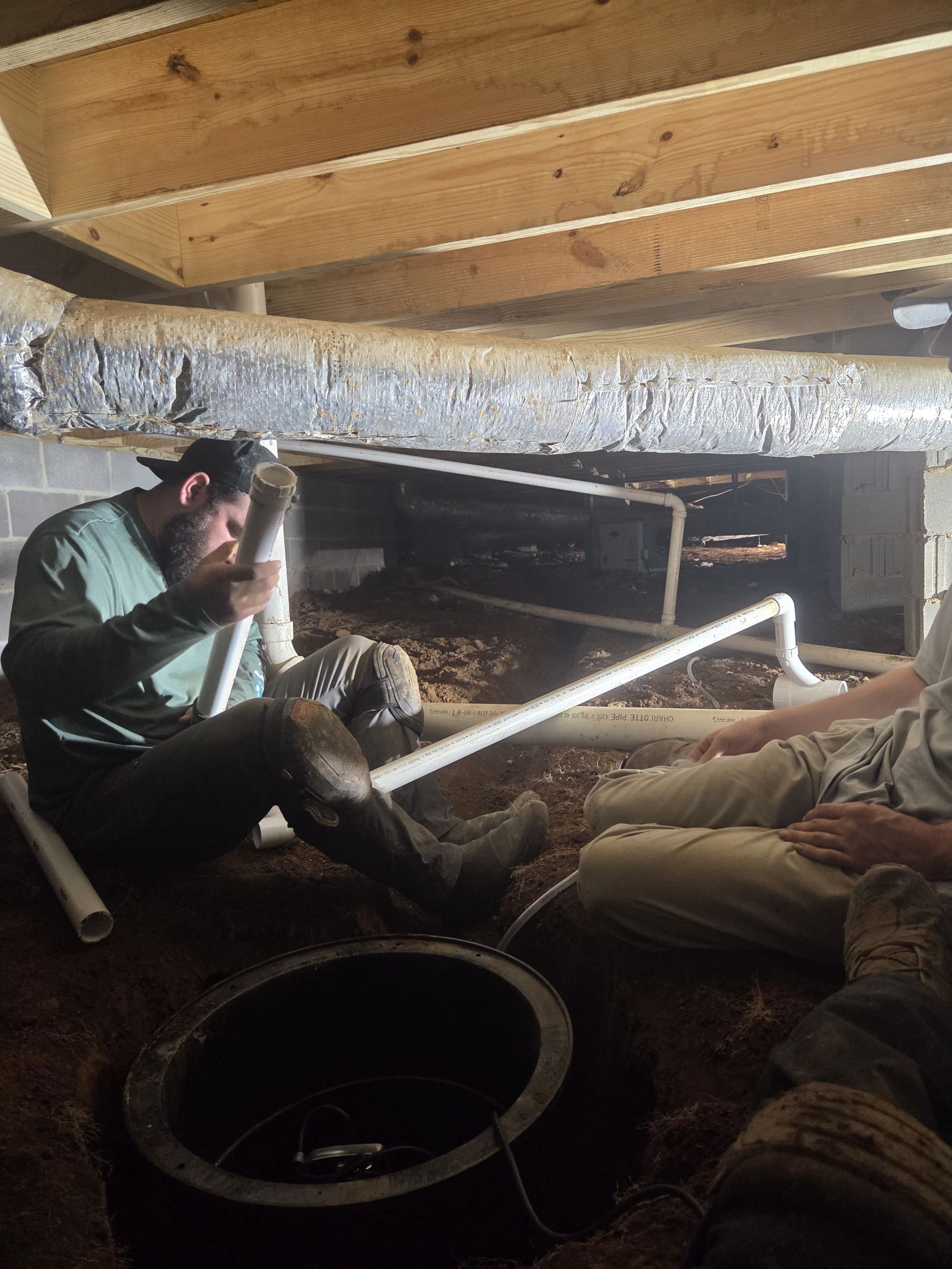 Crawlspace Cowboys crew installing a sump pump basin and PVC drainage piping inside a crawlspace